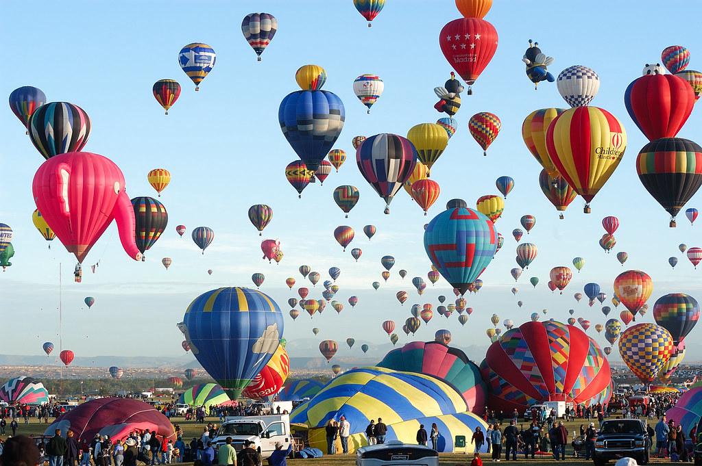 A stunning view of the Albuquerque Balloon Fiesta mass ascension at sunrise, with hundreds of balloons rising above the landscape and the Sandia Mountains in the background
