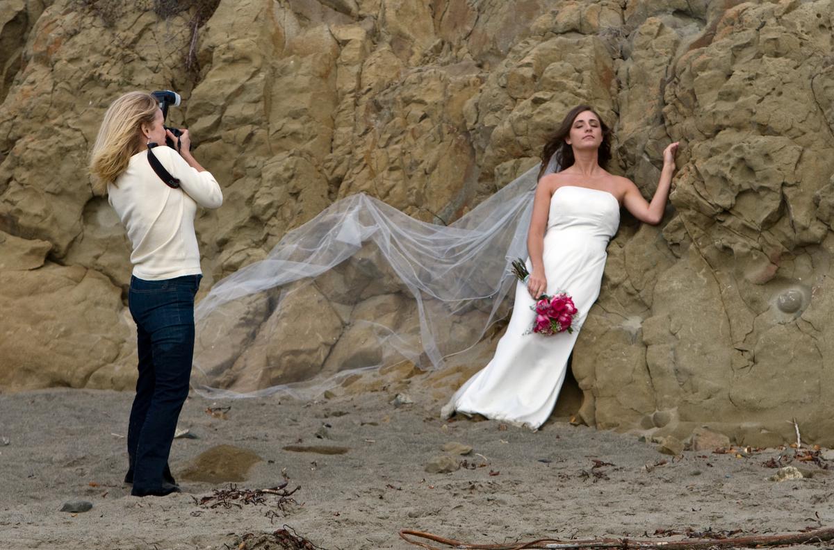 A candid photograph of Emily, a San Francisco-based wedding photographer, capturing a tender moment between a bride and groom in a natural, outdoor setting. Emily's focus and dedication to her craft are evident in her expression and the way she handles her camera.